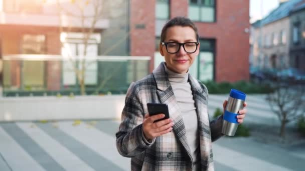 Retrato de una joven mujer de negocios caucásica con gafas y abrigo pasea por el distrito de negocios, con termo taza y usando smartphone. Comunicación, día de trabajo, concepto de vida ocupada. Movimiento lento — Vídeo de stock