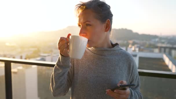 Femme commence sa journée avec une tasse de thé ou de café et vérifier les e-mails dans son smartphone sur le balcon à l'aube. — Video