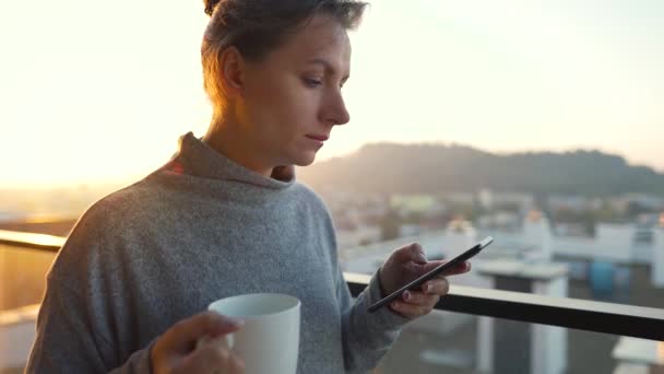 La mujer comienza el día con una taza de té o café y revisando correos electrónicos en su teléfono inteligente en el balcón al amanecer, en cámara lenta. — Vídeo de stock