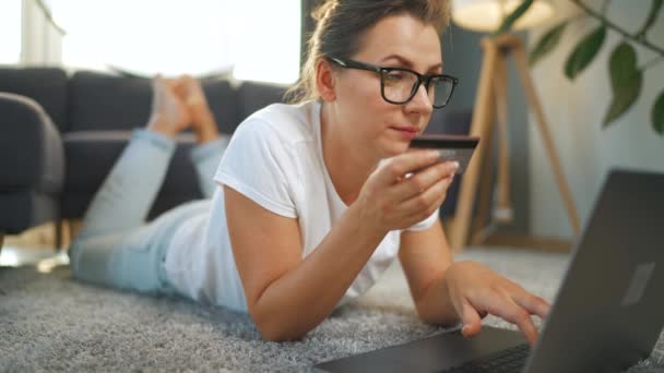 Kobieta w okularach leży na podłodze i dokonuje zakupu online za pomocą karty kredytowej i laptopa. Czarny kot przechodzi przez sofę i leży za nią. Zakupy online, technologia stylu życia — Wideo stockowe