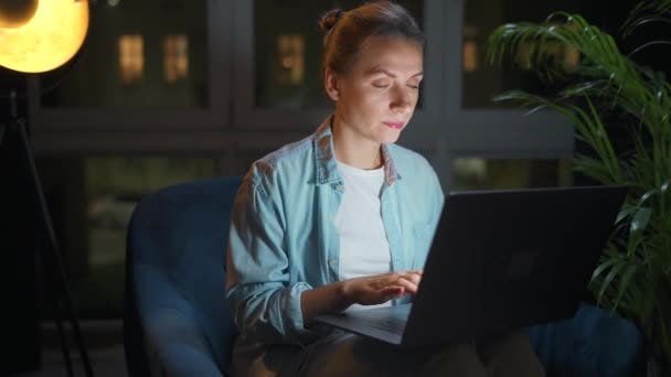 La mujer está sentada en el sillón y trabajando en un portátil por la noche. Concepto de trabajo remoto. — Vídeo de stock
