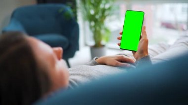 Kadın evde kanepede yatıyor ve dikey modda yeşil maket ekranlı akıllı telefon kullanıyor. Kız internette geziniyor, içeriği, videoları, blogları izliyor.