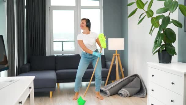 Mujer con auriculares limpiando la casa y divirtiéndose bailando con una escoba y un paño. Movimiento lento — Vídeo de stock