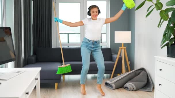 Kobieta w słuchawkach sprząta dom i dobrze się bawi tańcząc z miotłą i myjką. Zwolniony ruch — Wideo stockowe
