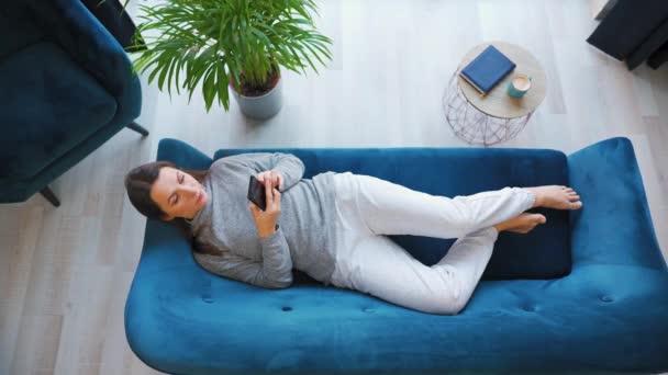 Overhead shot of happy relaxed woman holding smart phone, using mobile apps, watching funny video, having fun chatting in social media, lying on couch at home. — Stockvideo