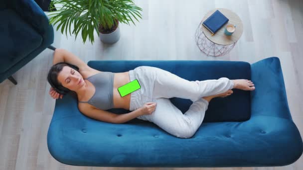 Nad głową zrelaksowana kobieta słuchająca wykładu lub muzyki w słuchawkach na smartfonie z zielonym ekranem makietowym, leżąca na kanapie w domu. — Wideo stockowe
