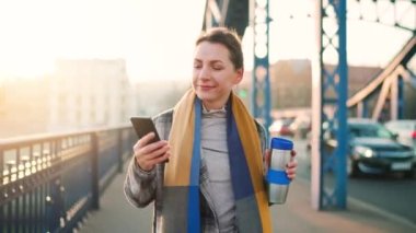 Paltolu beyaz bir iş kadınının portresi, soğuk bir sabahta köprüde yürüyor, kahve içiyor ve akıllı telefon kullanıyor. İletişim, iş günü, yoğun yaşam konsepti.
