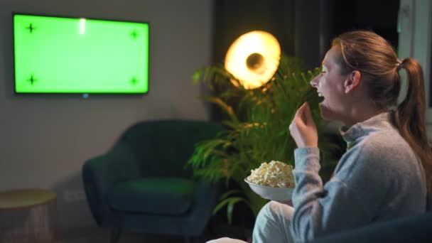 Vista posteriore di una donna seduta su un divano in soggiorno la sera e guardando un modello di schermo TV verde, si rallegra di ciò che vede.. — Video Stock