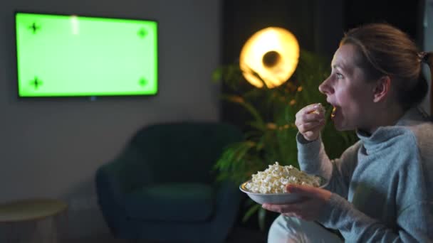 La donna seduta su un divano in salotto la sera e che guarda un mockup sullo schermo della TV verde, è emotivamente preoccupata per ciò che vede. La donna è infatuata di guardare e mangiare popcorn. — Video Stock