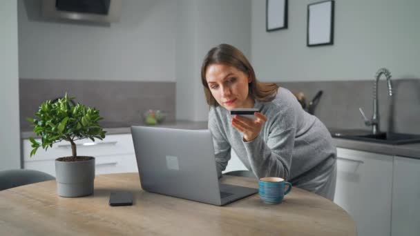 Kobieta stojąca w domu przytulnej kuchni, pochylając się nad stołem i robi zakupy online za pomocą karty kredytowej i laptopa. — Wideo stockowe