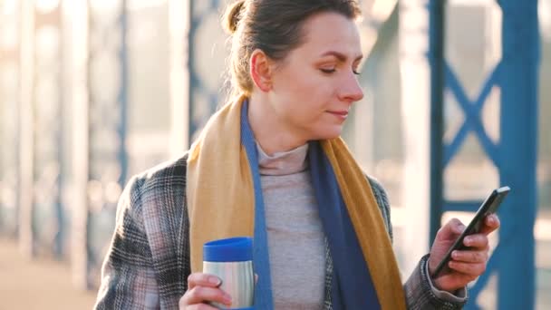 Ritratto di una giovane donna d'affari caucasica con un cappotto, che passeggia per la città in una mattinata gelida, beve caffè e usa lo smartphone. Comunicazione, giornata di lavoro, concetto di vita frenetica. Rallentatore — Video Stock