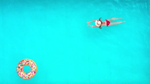 Θέα από την κορυφή ως γυναίκα με κόκκινο μαγιό κολυμπά στην πισίνα. Πολύχρωμα φουσκωτά ντόνατ επιπλέουν κοντά της. Θερινός τρόπος ζωής — Αρχείο Βίντεο
