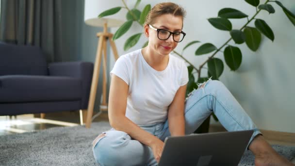 Woman with glasses is sitting on the floor and working on a laptop. Concept of remote work. — Stock Video