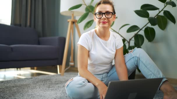 Portret van een vrouw met een bril die over een laptop naar de camera kijkt in het interieur van een gezellig appartement. Begrip werk op afstand. — Stockvideo