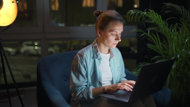 Donna è seduta sulla poltrona e lavora su un computer portatile di notte. Concetto di lavoro a distanza. — Video Stock