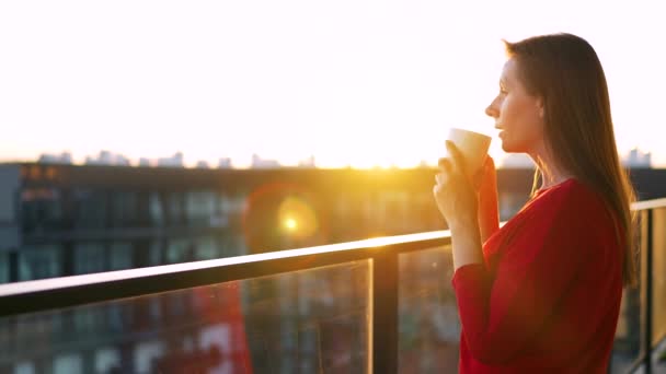 Kvinna i röd klänning med en kopp kaffe stående på balkongen och beundra solnedgången. Långsamma rörelser — Stockvideo