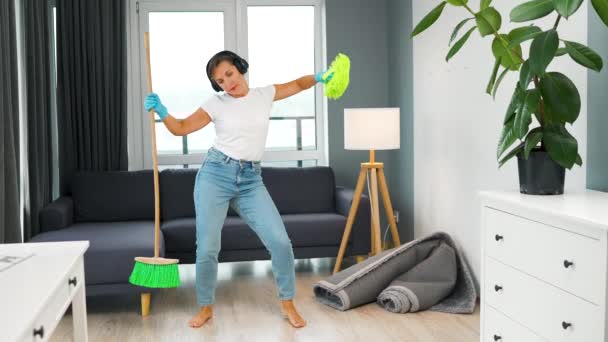 Mulher em fones de ouvido limpando a casa e se divertindo dançando com uma vassoura e pano. Movimento lento — Vídeo de Stock