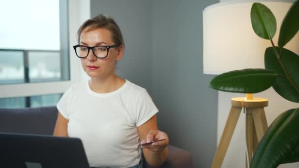 Vrouw met bril zit op een bank in een gezellige kamer en maakt online boodschappen met een creditcard en laptop. Online winkelen, lifestyle technologie — Stockvideo