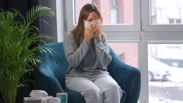 Una mujer malsana se sienta en una silla y estornuda o se sopla la nariz en una servilleta porque tiene un resfriado, gripe, coronavirus. Está nevando afuera. — Vídeos de Stock