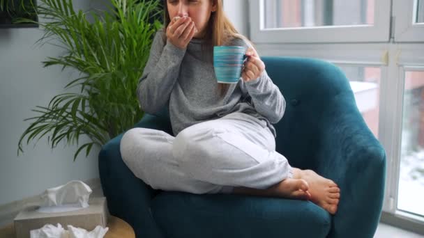 Niezdrowa kobieta siedzi na krześle, pije gorącą herbatę lub lek na przeziębienie i kicha lub wbija nos w serwetkę, bo ma przeziębienie, grypę, koronawirus. Na zewnątrz pada śnieg. — Wideo stockowe
