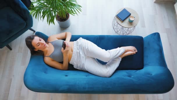 Vue aérienne d'une femme heureuse et détendue tenant un téléphone intelligent, utilisant des applications mobiles, regardant une vidéo drôle, s'amusant à bavarder dans les médias sociaux, allongé sur le canapé à la maison. — Video