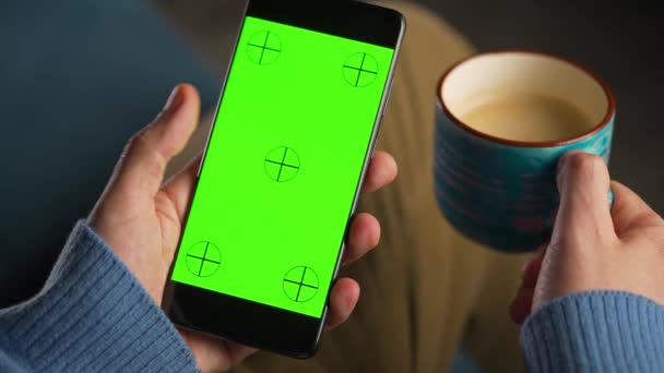 Mann benutzt Smartphone mit grünem Bildschirm im vertikalen Modus und trinkt Kaffee. Mann surft im Internet, schaut Inhalte, Videos. — Stockvideo