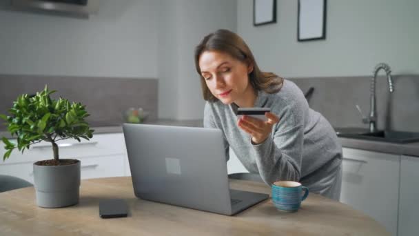 Žena stojící v domácí útulné kuchyni, naklání se přes stůl a dělá on-line nakupování pomocí kreditní karty a notebooku. — Stock video