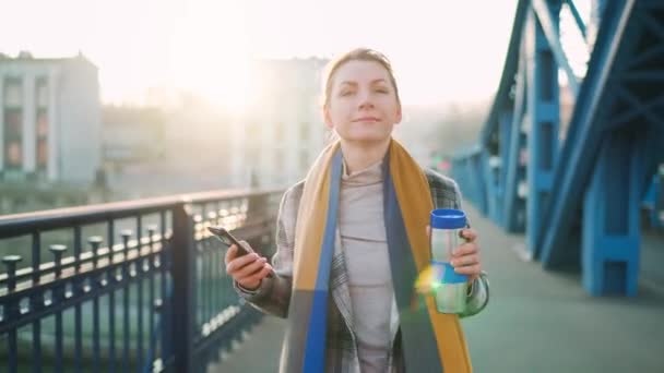 Retrato de una joven mujer de negocios caucásica en un abrigo, caminando por el puente en una mañana helada, bebiendo café y usando un teléfono inteligente. Comunicación, día de trabajo, concepto de vida ocupada. — Vídeo de stock