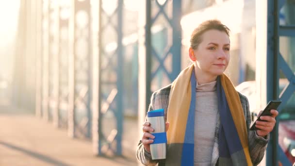 Portret młodej białej biznesmenki w płaszczu, spacerującej po mieście w mroźny poranek, pijącej kawę i korzystającej ze smartfona. Komunikacja, dzień pracy, pracowite życie. Zwolniony ruch — Wideo stockowe