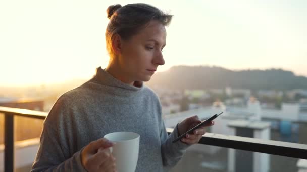 Frau beginnt ihren Tag mit einer Tasse Tee oder Kaffee und checkt im Morgengrauen auf dem Balkon E-Mails. — Stockvideo