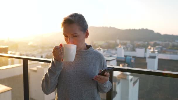 Femme commence sa journée avec une tasse de thé ou de café et vérifier les e-mails dans son smartphone sur le balcon à l'aube. — Video