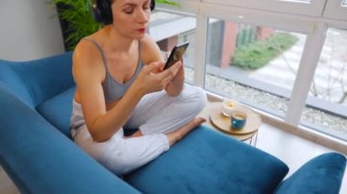 Kablosuz kulaklık takan, müzik dinleyen ve cep telefonu uygulamaları kullanan ya da akıllı telefonlu bir kadın evdeki kanepede oturuyor.