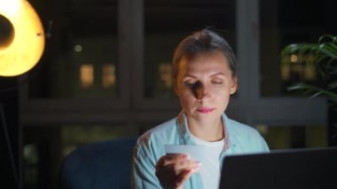 Kadın koltukta oturuyor ve geceleri kredi kartı ve dizüstü bilgisayar kullanarak online alışveriş yapıyor..