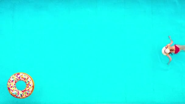 Blick von oben, als eine Frau im roten Badeanzug im Pool schwimmt. Bunte aufblasbare Donuts schweben in ihrer Nähe. Sommerlicher Lebensstil — Stockvideo