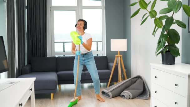 En kvinne med hodetelefoner som vasker huset og har det gøy med å danse med en kost og klut. Langsom bevegelse – stockvideo
