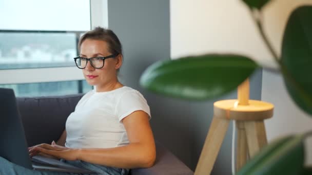 Žena s brýlemi sedí na gauči a pracuje na laptopu. Koncept práce na dálku. — Stock video