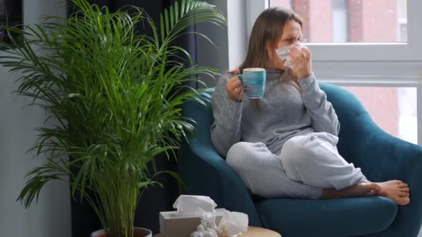 Niezdrowa kobieta siedzi na krześle, pije gorącą herbatę lub lek na przeziębienie i kicha lub wbija nos w serwetkę, bo ma przeziębienie, grypę, koronawirus. Na zewnątrz pada śnieg. — Wideo stockowe