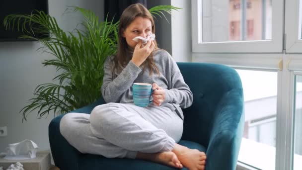 Donna malsana si siede su una sedia, beve tè caldo o una medicina fredda e starnutisce o soffia il naso in un tovagliolo perché ha un raffreddore, influenza, coronavirus. Fuori nevica. — Video Stock