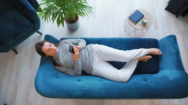 Nahoře záběr šťastné uvolněné ženy držící chytrý telefon, pomocí mobilních aplikací, sledování legračního videa, baví chatování na sociálních sítích, ležící na gauči doma. — Stock video