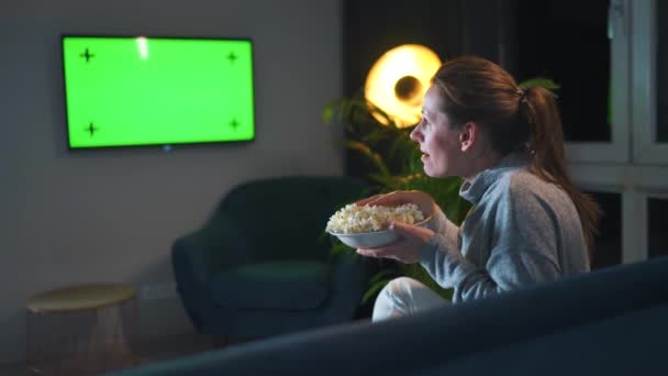 Vrouw die 's avonds in de woonkamer op een bank zit en naar een groene tv-scherm kijkt, maakt zich emotioneel zorgen over wat ze ziet. De vrouw is smoorverliefd op popcorn kijken en eten. — Stockvideo