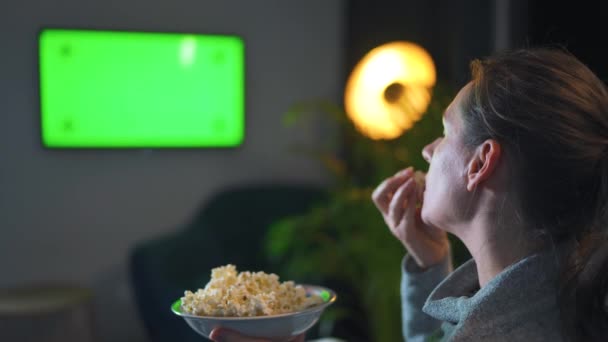 Vrouw zittend op een bank in de woonkamer in de avond en kijkend naar een groene TV scherm mockup. De vrouw is smoorverliefd op popcorn kijken en eten. — Stockvideo