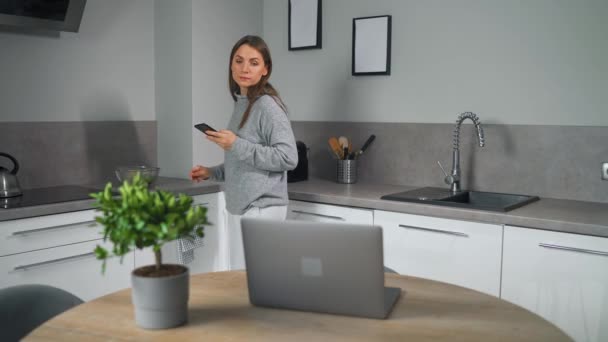 Femme avec smartphone à la main debout dans la cuisine confortable à la maison, se fait café et utiliser un ordinateur portable faire du travail indépendant. Concept de télétravail. Bureau à domicile — Video