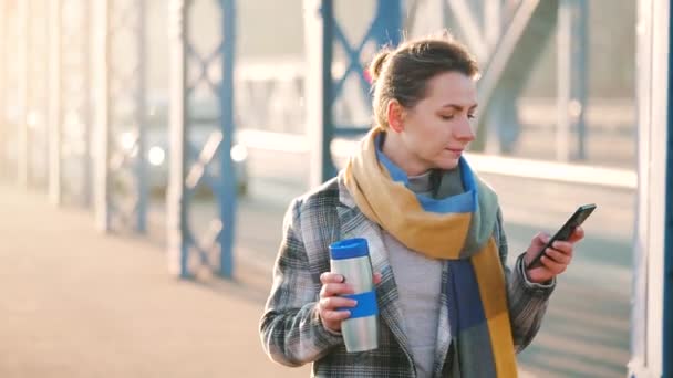 Portrait of a young caucasian businesswoman in a coat, walking around the city on a frosty morning, drinking coffee and using smartphone. Communication, work day, busy life concept. — Stock Video