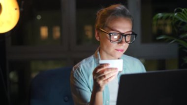 Kadın koltukta oturuyor ve geceleri kredi kartı ve dizüstü bilgisayar kullanarak online alışveriş yapıyor..