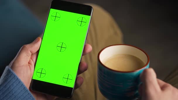 Adam yeşil ekran ekranlı akıllı telefonu dikey modda kullanıyor ve kahve içiyor. İnternette gezinen, içeriği izleyen, videolar. — Stok video