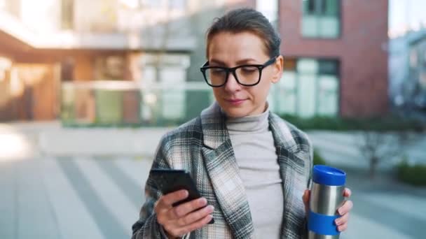 Ritratto di una giovane donna d'affari caucasica con occhiali e cappotto passeggia per il quartiere degli affari, con thermo cup e utilizzando lo smartphone. Comunicazione, giornata lavorativa, concetto di vita frenetica — Video Stock