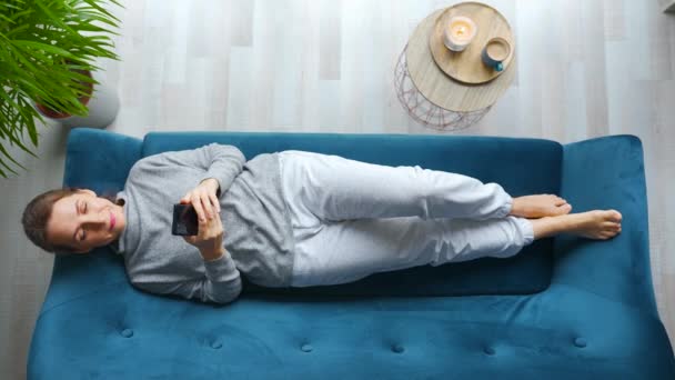 Fotografia aérea de mulher relaxada feliz segurando telefone inteligente, usando aplicativos móveis, assistindo vídeo engraçado, se divertindo conversando nas mídias sociais, deitado no sofá em casa. — Vídeo de Stock