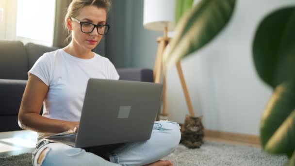 Gözlüklü kadın yerde oturuyor ve bir dizüstü bilgisayar üzerinde çalışıyor. Kedi arka planda yatıyor. Uzak çalışma kavramı. — Stok video