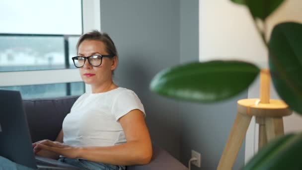 眼鏡をかけた女性がソファに座ってノートパソコンで働いている。リモートワークの概念. — ストック動画
