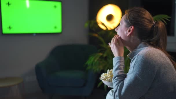 夜のリビングルームでソファに座って緑のテレビのモックアップを見ている女性のリアビューは、彼女が見ているものについて喜びです. — ストック動画